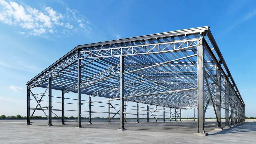 Çelik hangarın taşıyıcı iskeleti: cıvatalı kolon ayakları, kafes çatı makasları, aşıklar ve çapraz bağlantılar; beton döşeme dökülmüş, cephe kaplaması henüz yok.